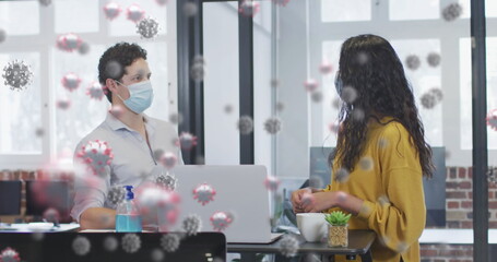 Image of covid 19 cells floating over man and woman wearing face masks, touching elbows in office
