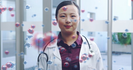 Asian female doctor smiles behind a digital composite of Covid-19 cells.