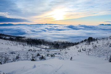 Fototapeta na wymiar 雪に覆われた山の斜面と眼下の雲海の雄大な冬景色。雲の空の朝陽。北海道の美幌峠の朝。