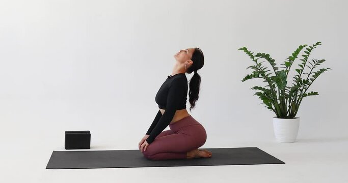 A brunette performs deep bends, extension and flexion of the spine in vajrasana, training in a bright room in sportswear, sitting on a mat with her legs tucked under her