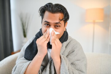 Sick man sitting at home on sofa in living room, sneezing has allergy and runny nose, Indian man with napkin has a cold