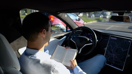 Male businessperson reading book during riding on electrical vehicle with autopilot at urban road. Successful businessman improving his knowledge while riding an autonomous self driving electric car - 757921052