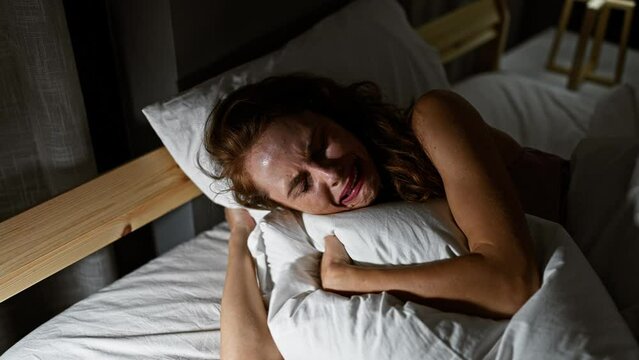 A distraught young woman embracing a pillow while lying in bed indoors, conveying a sense of sadness or heartache in a dimly lit bedroom setting.