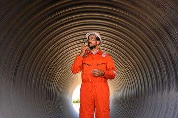 An engineer in a reflective orange jumpsuit and hard hat stands inside a corrugated metal tunnel using a walkie-talkie..