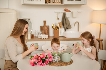 Family communication of mom and children. Happy mother with her daughter and son drinking tea or cocoa in the kitchen and having fun. Children congratulated their mother with flowers on Mother's Day.