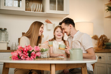 Obraz na płótnie Canvas Mother, father, and daughter drinking tea or cocoa sitting in the kitchen, and having fun. Family communication and spending time together. Mom and Dad hug a child. Mommy and Daddy embrace children.
