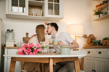 Fototapeta na wymiar Romantic female and male kissing, spending time together at home. Loving young man and woman having conversation and drink tea in morning. Couple drinking coffee, holding cups in hands in the kitchen.
