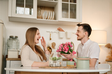 Obraz na płótnie Canvas Man congratulate woman and give bouquet of flowers on Women's day. Romantic female and male, spending time together. Loving young couple having conversation and drinking coffee in morning in kitchen.