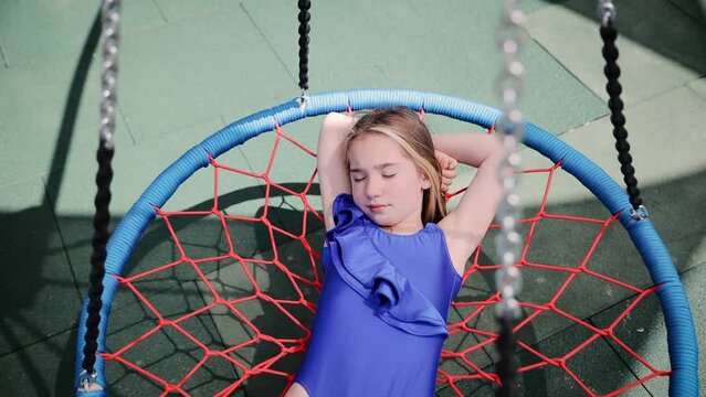 Portrait of adorable child lying and dreaming on net swing on child playground. Smiling little caucasian girl wearing blue swimsuit riding on swing with net outdoors near the pool
