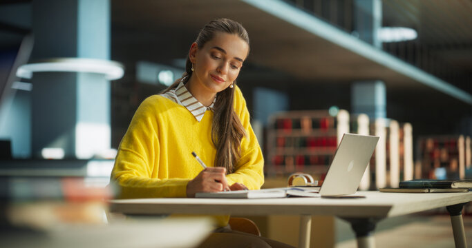 Thoughtful European College Student Using Laptop Computer to Study in a Public Library. Beautiful Young Woman Learning Online, Getting Ready for Exams, Taking Notes and Drafting an Essay