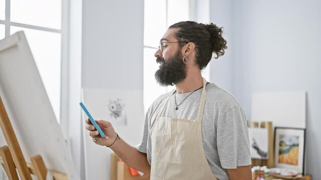 A bearded young man in an apron smiles while using a smartphone in a bright art studio.