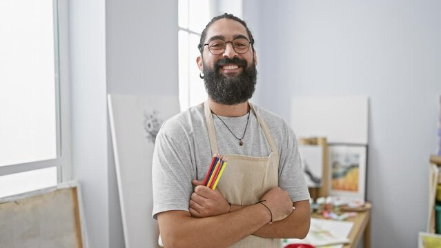 Smiling bearded young man holding colored pencils crosses arms in a bright studio setting.