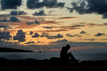 unrecognizable girl in silhouette sitting at the edge of steep and rugged cliffs of the gulf of biscay bay near St. Jean de Luz and using her smart phone with dramatic sunset and cloudscape.