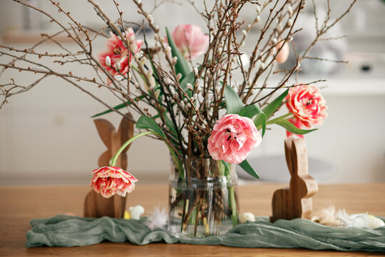 Stylish easter decoration on wooden table in modern farmhouse living room. Beautiful tulips bouquet with willow branches, easter eggs and wooden bunny figurines decor. Happy Easter!