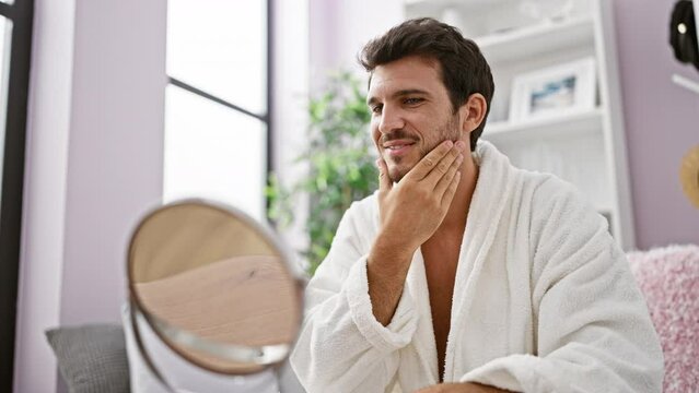 A handsome young man in a white bathrobe touches his beard, contemplating grooming in a cozy home interior.