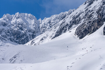 Trekking and skituring in High Tatra Mountains in winter. Trail in snow with people and the giant rocky mountains in the bacground in sunny day with clear blue sky.