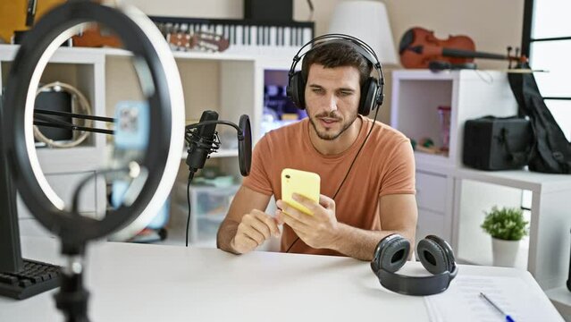Handsome hispanic man with beard using smartphone in music studio with microphone and headphones