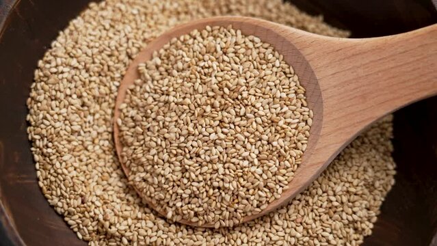 Sesame seasoning seeds falling on a wooden spoon in brown bowl. Culinary ingredients close up. Slow motion