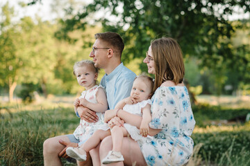 A happy family is sitting on bench on shore of lake at sunset. Mom, dad hug children daughter in the summer park. A loving couple with two childs. Mother, father embrace kids and spend time together.