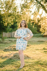 Fototapeta premium A beautiful woman in glasses stands on green grass at sunset. Female portrait looking at a camera on a summer day. A happy girl in a dress on the field in the sunset background.