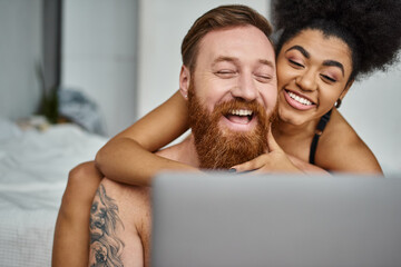 tattooed man and african american woman watching comedy movie on laptop and laughing together