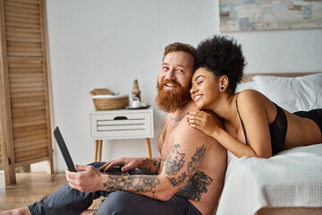 bearded man and african american woman, interracial couple lying on bed and watching movie on laptop - 757968079