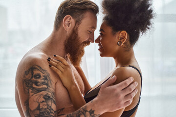 happy bearded and tattooed man without shirt hugging his african american girlfriend near curtain