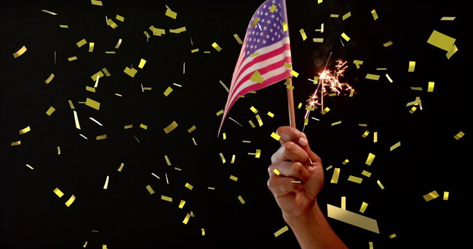 Image of confetti falling over hand holding flag of united states of america and sparkler