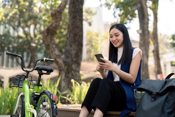 Asian businesswoman with bicycle using smartphone and sitting outside the office building. Woman commuting on bike go to work. Eco friendly vehicle, sustainable lifestyle concept
