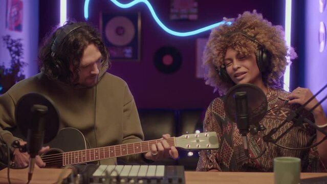 UCC of energetic young Black lady recording hip-hop song and Caucasian man accompanying her on guitar at neon lit home studio at night