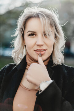Stylish Woman in Black Coat with Blonde Hair and Watch, Gazing at Camera with Thoughtful Gesture. A woman with wearing a black coat looks into the camera. She holds her thumb near her mouth