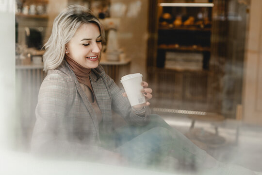 A woman is sitting in a cafe with a cup of coffee in her hand. She is smiling and she is enjoying her time. Cafe Bliss. Smiling Woman Savoring Coffee in Cozy Atmosphere.