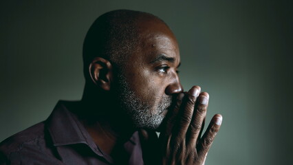 One religious black senior man in contemplative PRAYER at home in dimly lit room with eyes closed having HOPE and FAITH during challenging times. Spiritual African American 50s person - obrazy, fototapety, plakaty