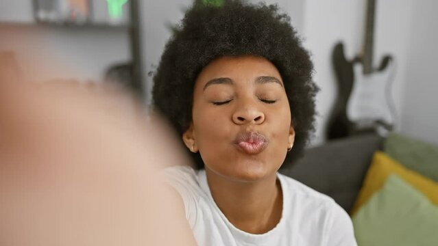 A joyful woman with afro hair smiles and makes a kissy face for a selfie in a cozy living room, showcasing positivity and lifestyle.