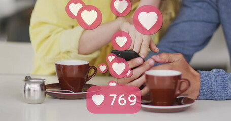 Image of heart icons floating over caucasian couple using smartphone and drinking coffee