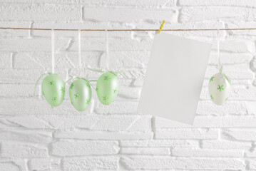Green Easter eggs and blank card mockup hanging on a rope, white brick background. Side view, selective focus. - 757985843