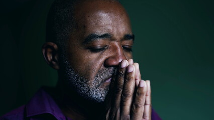 Spiritual African American senior person praying to GOD, closing eyes in contemplation with hands clenched together. Faithful man asking for HELP and SUPPORT - obrazy, fototapety, plakaty