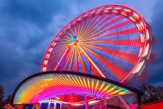 Colorful Ferris wheel at a funfair in Germany with motion blur. Glowing red, yellow lines and circles from long exposure light trails. Popular twilight ride at blue hour. Roundabout or merry-go-round. - Powered by Adobe