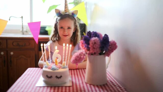 Happy little girl with unicorn hair decoration celebrating her sixth birthday, blowing on candles and making a wish.