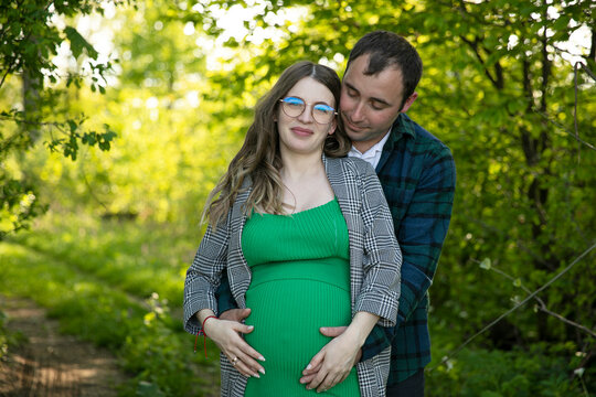 Front image of a happy couple expecting a baby spend time outdoors on a sunny day, being hugged. Horizontal view.