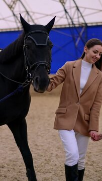 Lady and her little daughter attending horse riding lessons at drill-hall. Woman is holding a beautiful black horse by the bridle. Vertical video.
