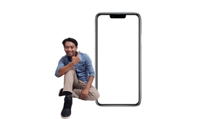 Full size asian young man in blue shirt sitting near big blank screen mobile phone with mockup area using smart phone isolated on plain white background