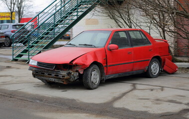 A red wrecked car stands near a white brick wall of a house, Podvoysky Street, St. Petersburg,...