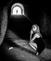 A beautiful blonde woman with perfect legs sits in the old brick tunnel near the stairs and a paper airplane with the man's silhouette upstairs. Black and white retro conceptual photo.