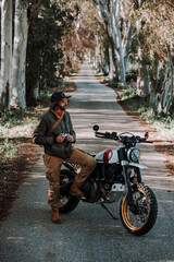 Chico con barba con atuendo de motorista posando con su moto y su cámara 