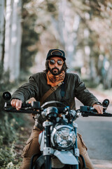Chico con barba con atuendo de motorista posando con su moto y su cámara 