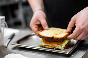 A close-up view of hands assembling a freshly made sandwich with toasted bread, lettuce, and tomatoes in a professional kitchen setting