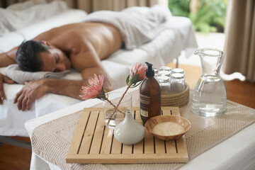 Salt, flowers and man in spa to relax on bed or break with luxury pamper treatment tools on table. Protea, facial oil or person at resort and salon for natural healing benefits or massage with water