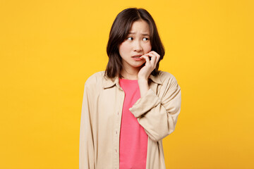 Young confused puzzled sad woman of Asian ethnicity wear pink t-shirt beige shirt pastel casual clothes look aside biting nails fingers isolated on plain yellow background studio. Lifestyle portrait.