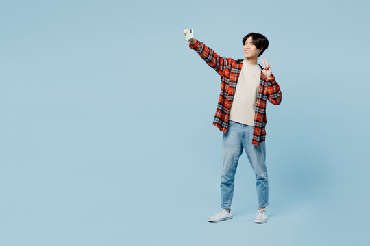 Full body young man of Asian ethnicity wears red hoody casual clothes do selfie shot on mobile cell phone post photo on social network show v-sign isolated on plain blue background. Lifestyle concept.